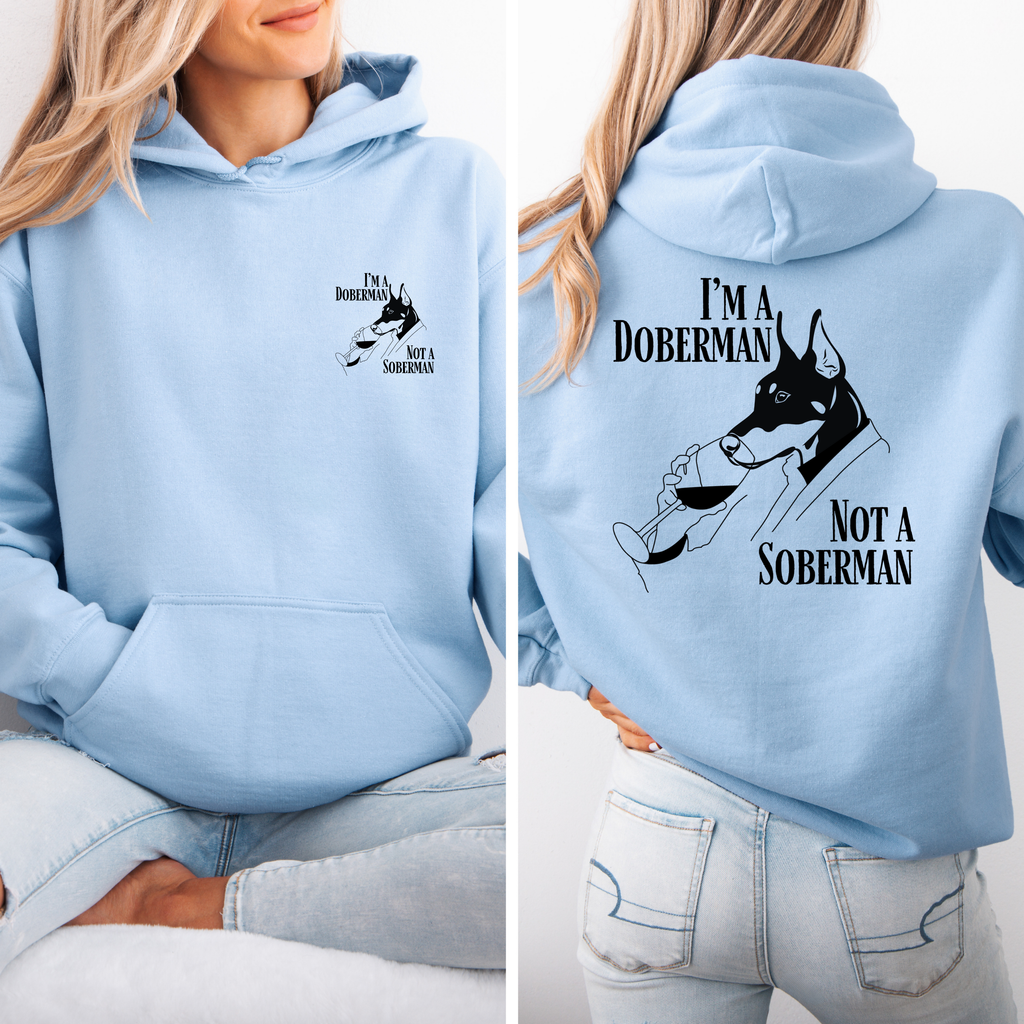 Hellblauer Hoodie mit kleinem Frontprint und großem Rückenmotiv „I’m a Doberman, Not a Soberman“ inklusive Dobermannzeichnung; Frau trägt den Hoodie im Sitzen.