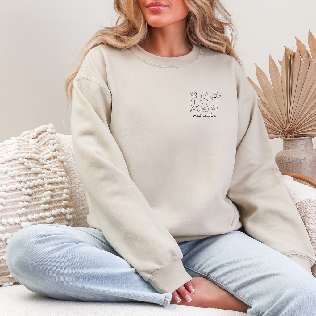 Beigefarbener Sweater mit Hunde-Yoga-Motiv und dem Schriftzug „Namaste“ als kleiner Brustprint, getragen von einer Frau im Sitzen.