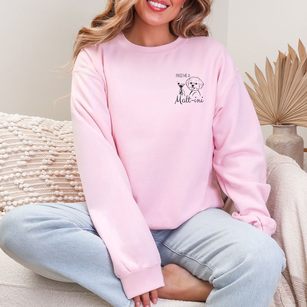 Rosafarbener Sweater mit kleinem Brustmotiv eines Maltesers mit Cocktailglas und dem Text „Pass me a Malt-ini“, getragen von einer sitzenden Frau.